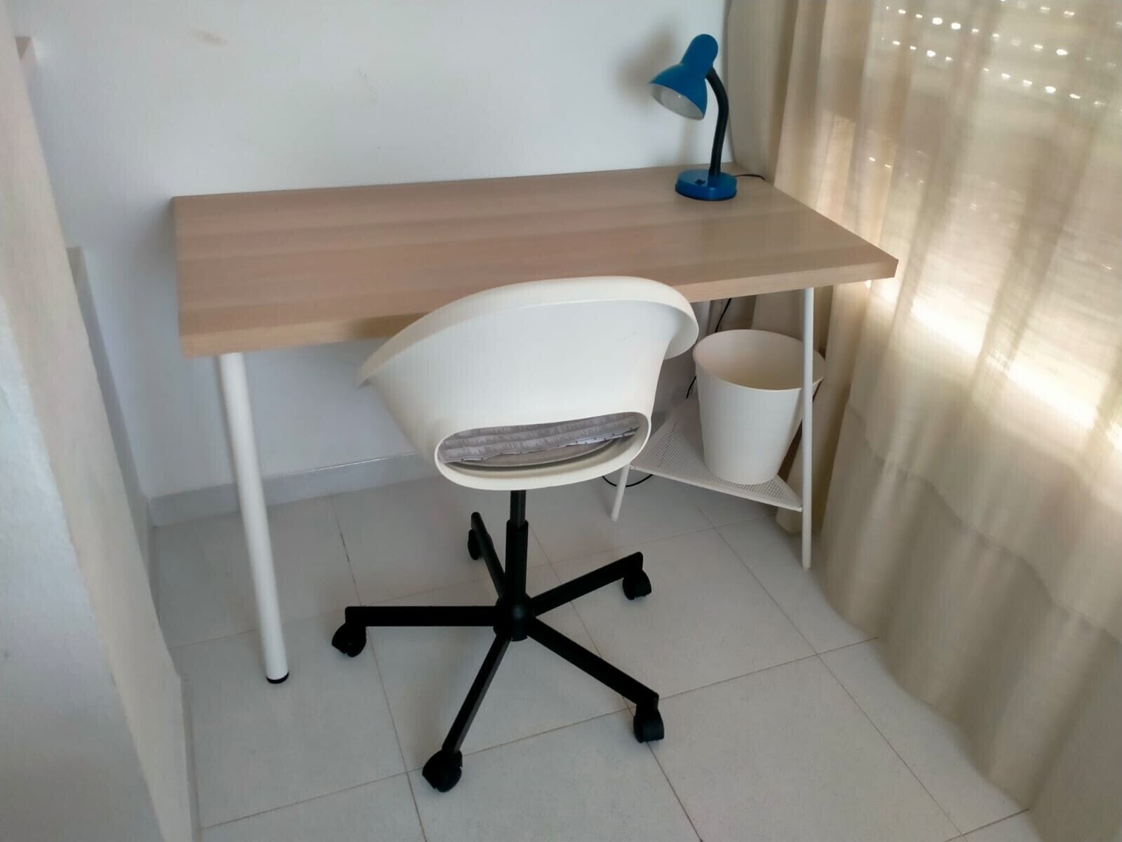 Een kleine kantoorruimte met een houten bureau, een witte stoel, een blauwe bureaulamp en een witte prullenbak.