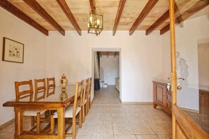A dining area with wooden chairs and a table, a hanging light fixture, and a view into another room through an open doorway.