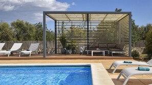 Un espace aménagé au bord de la piscine, comprenant un coin salon couvert, des chaises longues et une petite table agrémentée d’un vase de fleurs.