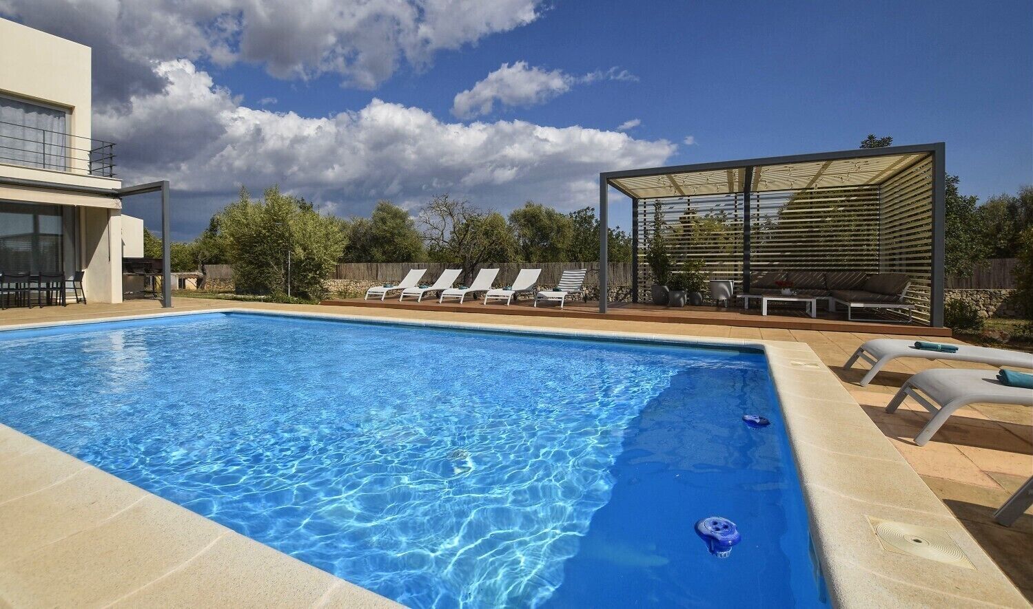Une piscine rectangulaire à l’eau d’un bleu limpide, entourée d’une terrasse carrelée. On y trouve des chaises longues et un espace de détente couvert, surmonté d’un toit en métal.