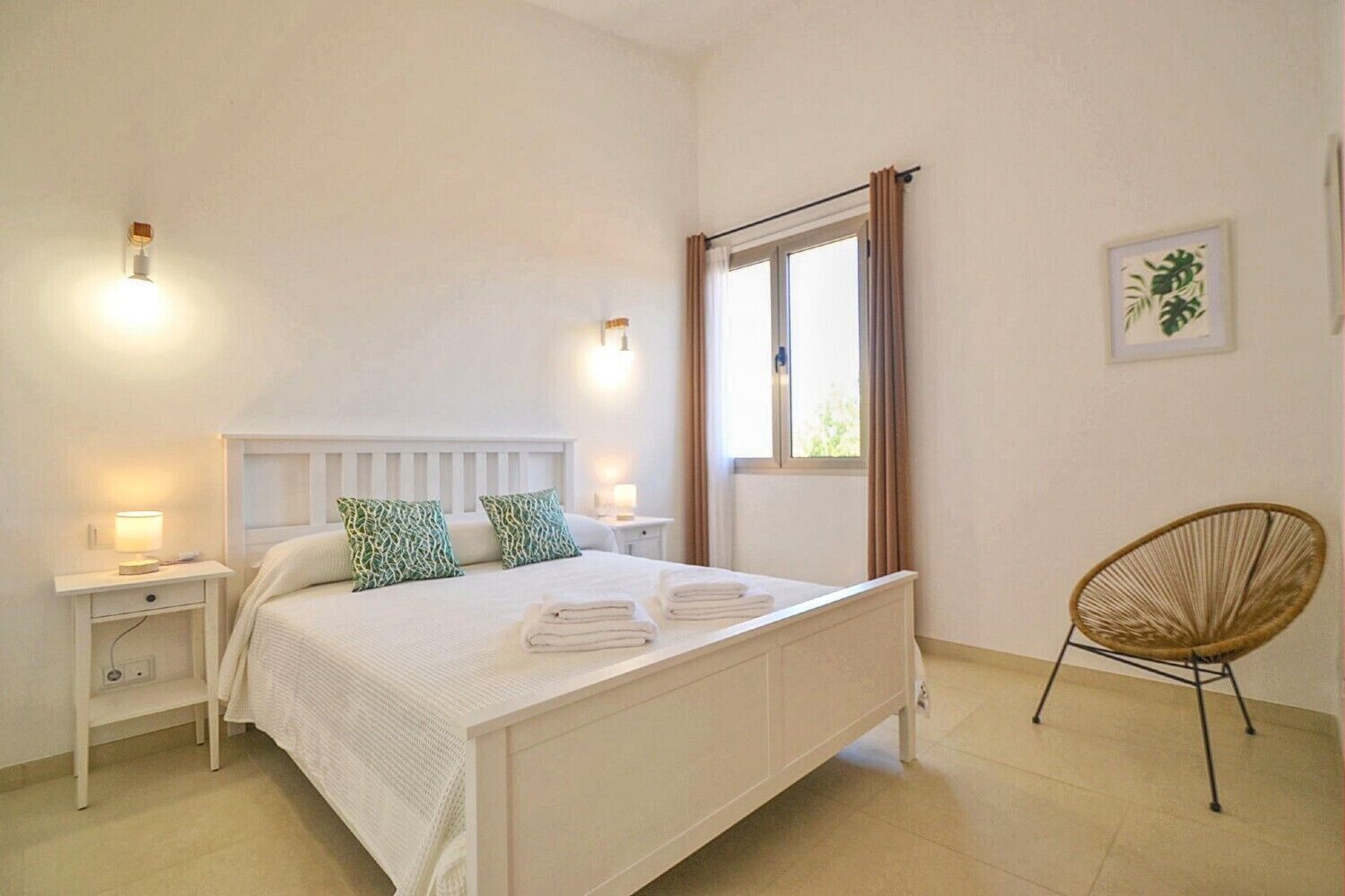 Un dormitori amb un llit, taules de nit, una cadira, una finestra amb cortines i llums penjades a la paret.