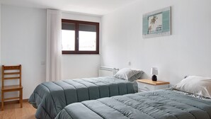 Camera da letto con due letti, una sedia di legno, un comodino con una lampada, una finestra con tende e un quadro appeso al muro.