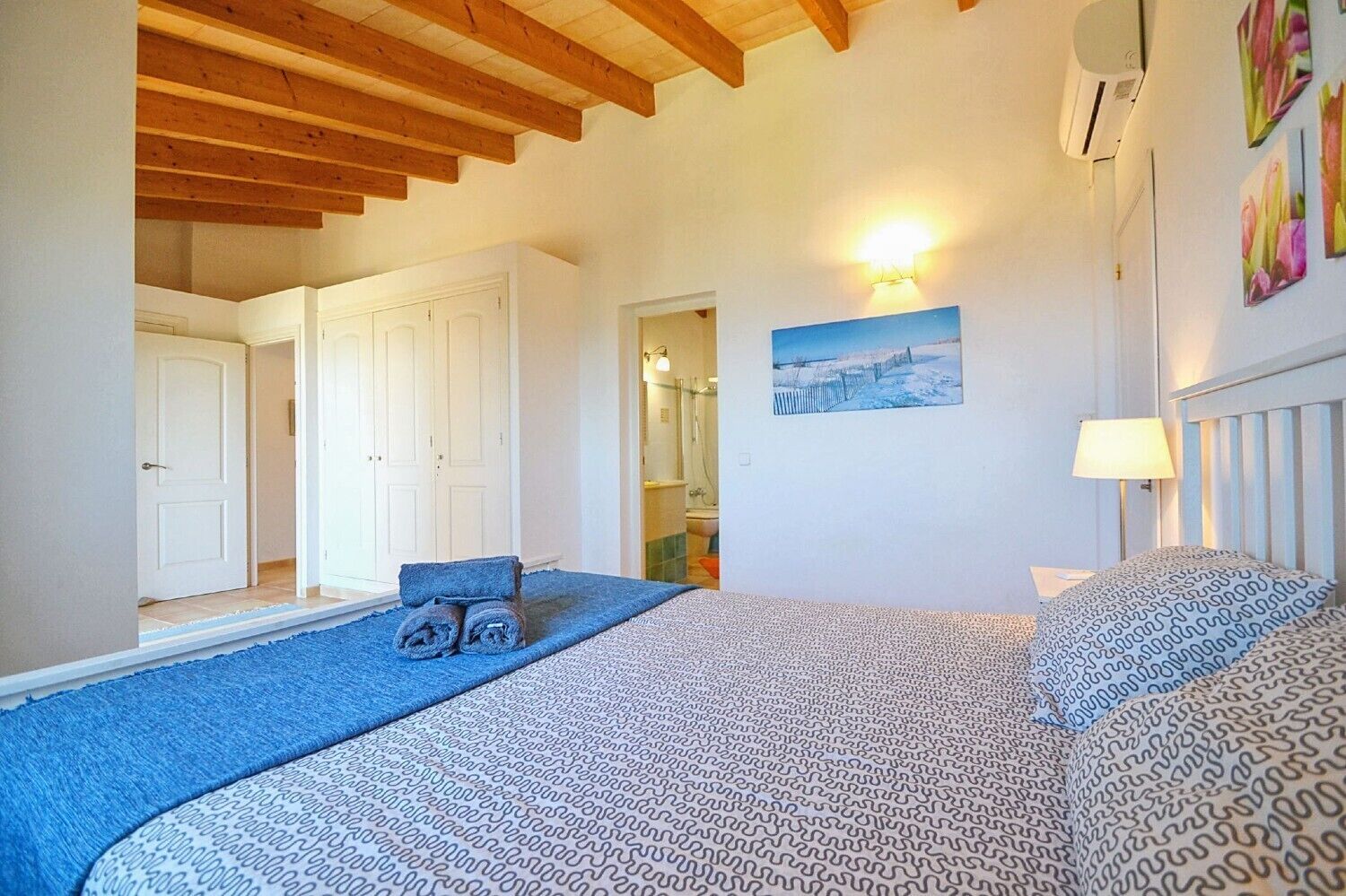 A bedroom with a bed, a blue area rug, a wall-mounted lamp, and a painting.