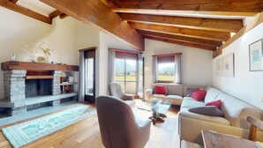 A cozy living room with a stone fireplace, wooden ceiling beams, a sofa set, and a view of the outdoors. - 4-room apartment in the loft. (Sils/Segl Maria)