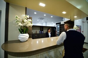 A reception area with a marble counter, a vase of white orchids, and a woman standing behind the counter. - HOTEL EBOLI (Eboli)