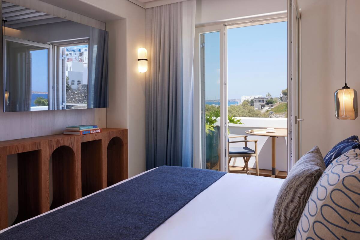 Habitación de hotel con una cama, una consola de madera con libros, un ventanal con cortinas y vistas al exterior.