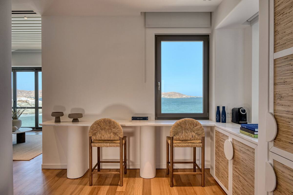 Una cocina moderna con un amplio ventanal con vistas al mar, dos taburetes de bar y una encimera blanca con algunos objetos.