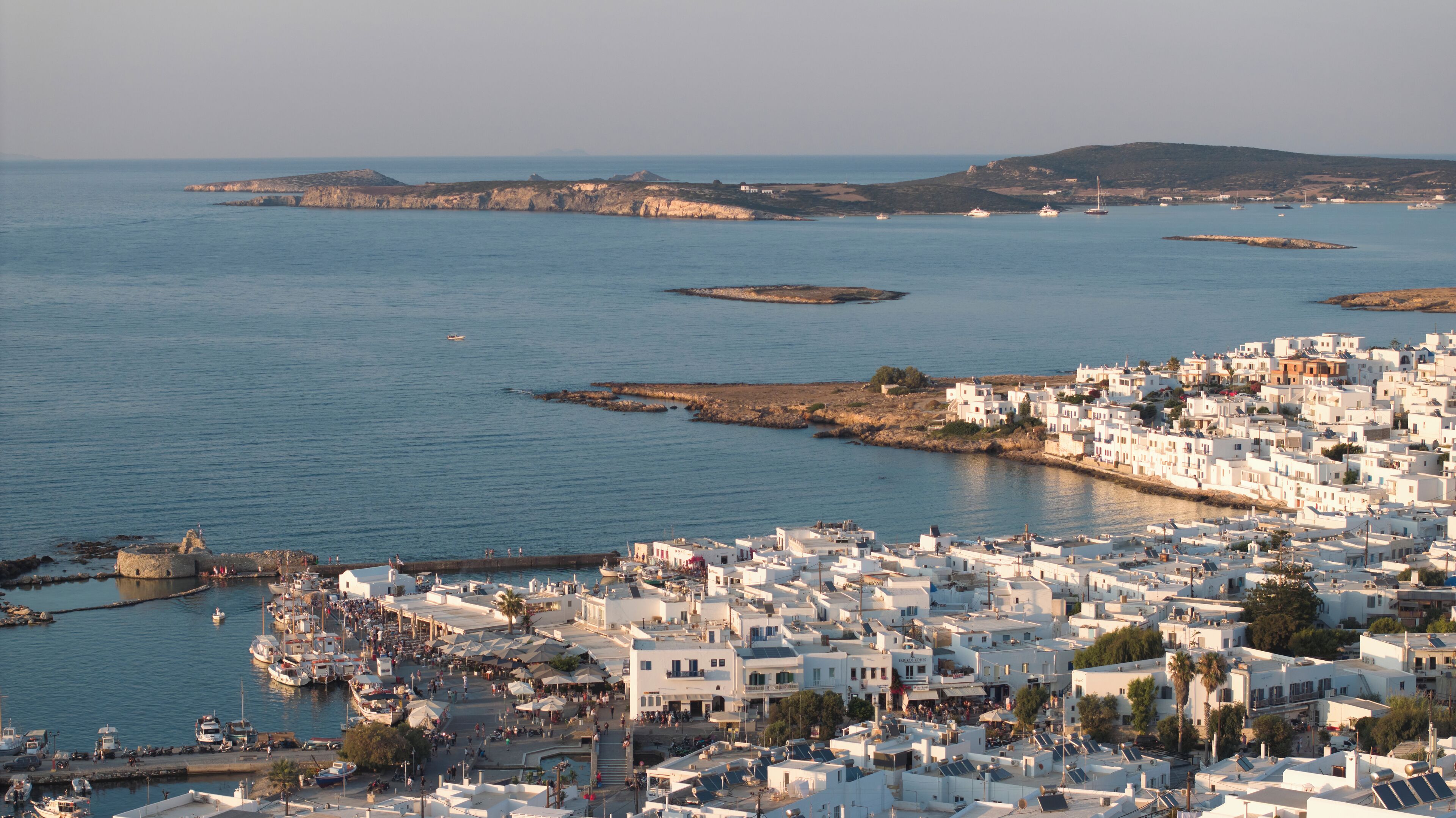 A coastal town with white buildings, a harbor with boats, and rocky outcrops in the background.
