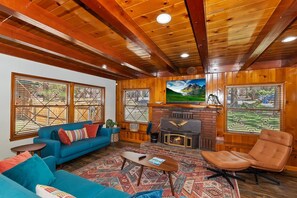 Una sala de estar acogedora con un sofá azul, un sillón color arena, una chimenea y grandes ventanales con vistas a un paisaje montañoso.