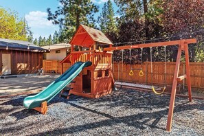 A wooden play structure with a slide and swings in a backyard. - Wren's Cottage by Destination Big Bear: Pet Friendly! Spa! Located in the Summit Lake Neighborhood! Infinity Game Table! Fenced Yard! Playgym! Horseshoe Pit! (Big Bear Lake)