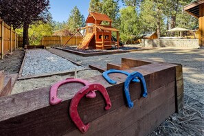 A playground with a wooden slide and climbing structures, surrounded by a wooden fence and trees. - Wren's Cottage by Destination Big Bear: Pet Friendly! Spa! Located in the Summit Lake Neighborhood! Infinity Game Table! Fenced Yard! Playgym! Horseshoe Pit! (Big Bear Lake)