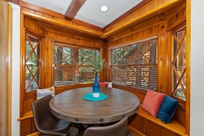 Un interior de madera con una mesa redonda y sillas, ventanas amplias con travesaños y vistas al exterior.