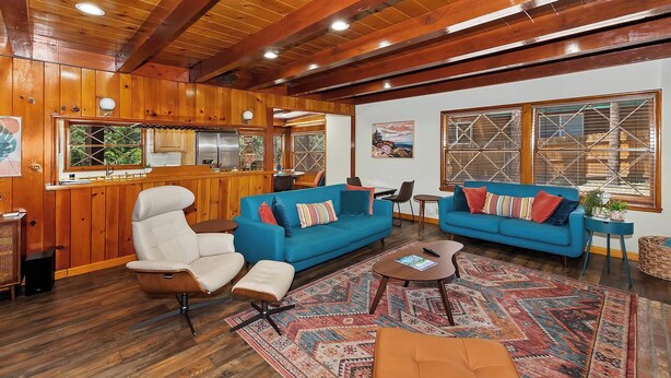 Interior acolhedor de uma cabana com paredes e teto de madeira, uma cozinha compacta, dois sofás azuis, uma poltrona branca e um tapete estampado.