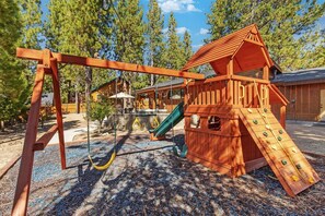 A wooden play structure with a slide and swings in a wooded area. - Wren's Cottage by Destination Big Bear: Pet Friendly! Spa! Located in the Summit Lake Neighborhood! Infinity Game Table! Fenced Yard! Playgym! Horseshoe Pit! (Big Bear Lake)