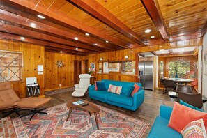 A spacious living area with a wooden ceiling, a blue sofa, a brown leather chair, and a dining table with chairs. - Wren's Cottage by Destination Big Bear: Pet Friendly! Spa! Located in the Summit Lake Neighborhood! Infinity Game Table! Fenced Yard! Playgym! Horseshoe Pit! (Big Bear Lake)