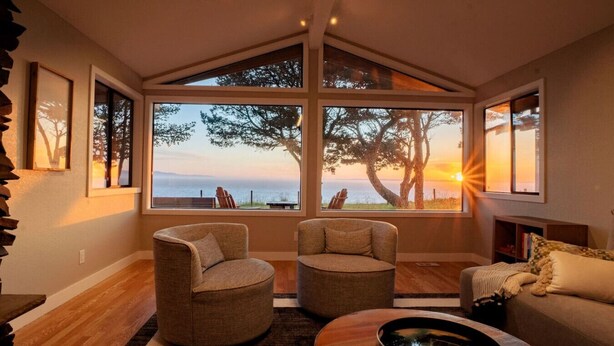 A modern living room with a large window offering a scenic view of trees and a sunset. There are two armchairs and a sofa arranged around a wooden coffee table. - Oceanfront Family Retreat (Oak Harbor)