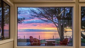 A room with a large window overlooking a sunset over a body of water, two Adirondack chairs, and a small table. - Oceanfront Family Retreat (Oak Harbor)