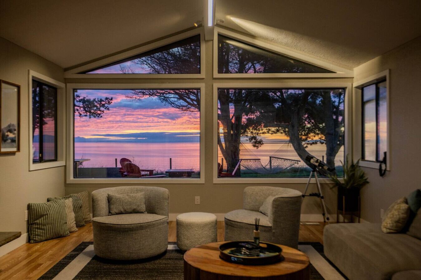 A living room with a large window overlooking a body of water, a hammock, and a view of a sunset.