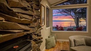 A cozy room with a stone wall, a fireplace, a large window with a sunset view, and a comfortable seating area. - Oceanfront Family Retreat (Oak Harbor)
