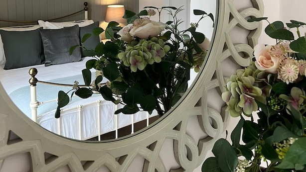 Un dormitorio con cama, mesita de noche, lámpara y un espejo decorativo con un arreglo floral.