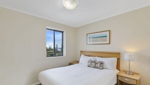 Une chambre à coucher avec un lit, une table de chevet, une lampe, une fenêtre et un tableau encadré au mur.