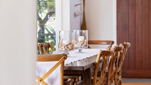 Une table à manger en bois, recouverte d’une nappe blanche, est entourée de chaises en bois. Deux chandeliers en verre, ornés de coquillages et contenant des bougies, sont posés sur la table.