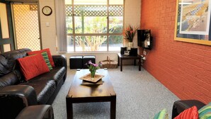 A living room with a black leather sofa, a coffee table with a vase of flowers, a TV on a stand, and a window with a view of greenery. - Acacia @ Boomerang Beach (Boomerang Beach)