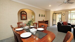 A dining area with a wooden table set for four, surrounded by chairs. There is a fireplace with a decorative arch above it, and a bookshelf with books and decorative items. The room has a cozy atmosphere with a couch and a window in the background. - Acacia @ Boomerang Beach (Boomerang Beach)