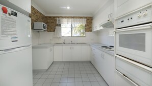 A compact kitchen with white cabinets, a microwave, a stove, and a window with a lace curtain. - Acacia @ Boomerang Beach (Boomerang Beach)