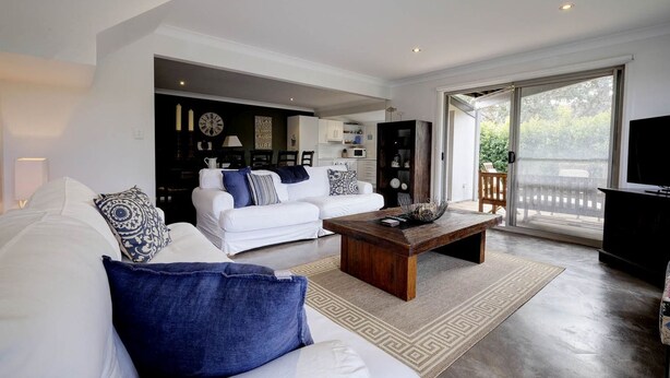 A modern living room with a white sofa, a coffee table, and a TV. There is a kitchen in the background with cabinets and a microwave.