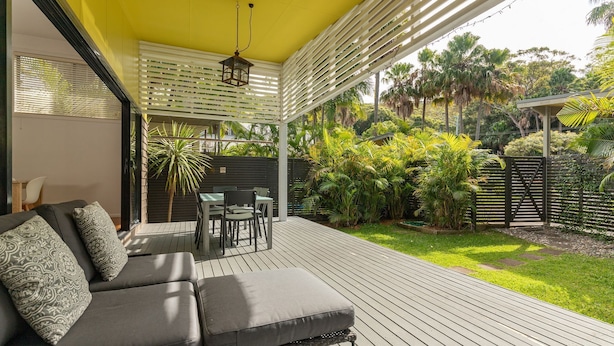 Un espacio al aire libre cubierto con un sofá y una mesa, rodeado de abundante vegetación y una terraza de madera.