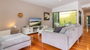 Una sala moderna con un sofá, una mesa de centro, un televisor sobre un mueble blanco y un ventanal con vistas a zonas verdes.