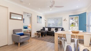 A modern living room with a grey sofa, a small armchair, a dining table with white chairs, and a ceiling fan.