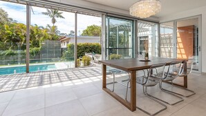 A modern dining area with a pool view, large sliding glass doors, and a wooden table. - Boof's at Blueys Beach (Blueys Beach)