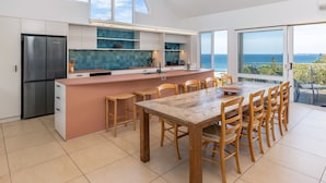 Une cuisine moderne dotée d’un îlot central, d’un coin repas avec des chaises en bois et offrant une vue sur la mer grâce à de grandes fenêtres.