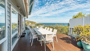 Une terrasse en bois avec une table et des chaises blanches, offrant une vue panoramique sur l’océan et les collines.
