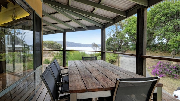 A wooden table and chairs under a covered patio with a view of a lake and mountains. - Far Shore @ Seal Rocks (Seal Rocks)