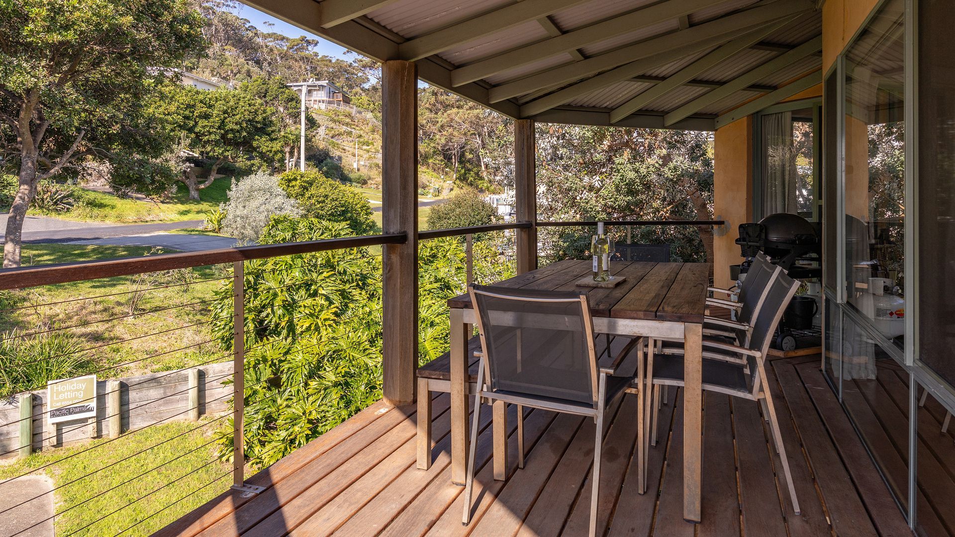 A wooden deck with a table and chairs, overlooking a landscaped area with trees and a road.