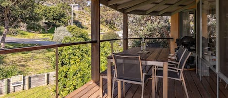 A wooden deck with a table and chairs, overlooking a landscaped area with trees and a road.