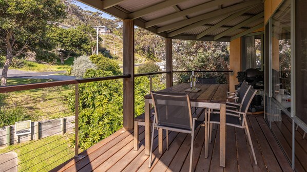 A wooden deck with a table and chairs, overlooking a landscaped area with trees and a road.