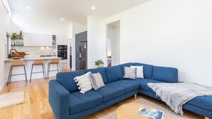 Ein modernes Wohnzimmer mit einem blauen Ecksofa, einer Küche mit weißen Schränken und einem Essbereich mit Barhocker.