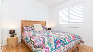 Un dormitorio con estructura de cama de madera, ropa de cama floral, dos lámparas de noche y una ventana con persianas.