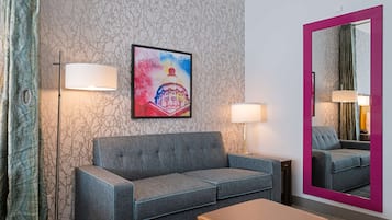 Une chambre d’hôtel avec un canapé gris, un petit pouf, deux lampadaires, un grand miroir et un mur recouvert d’un papier peint à motifs floraux.