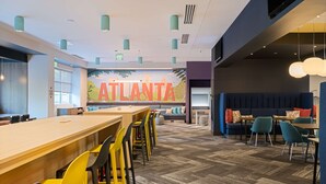 A modern interior with a long wooden table and yellow chairs, a blue sofa, and a mural with the word "ATLANTA". - Home2 Suites By Hilton Atlanta Airport College Park (College Park)