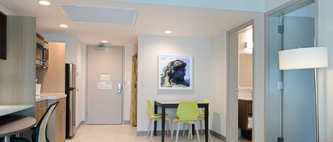 A modern hotel room with a kitchenette, a dining area with a table and chairs, a sofa, and a lamp.