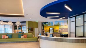 A hotel reception area with a curved counter, a green sofa with patterned cushions, and a mural on the wall depicting a cityscape. - Home2 Suites By Hilton Atlanta Airport College Park (College Park)