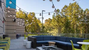 An outdoor patio with a blue sofa, yellow chairs, a fire pit, and string lights. - Home2 Suites By Hilton Atlanta Airport College Park (College Park)