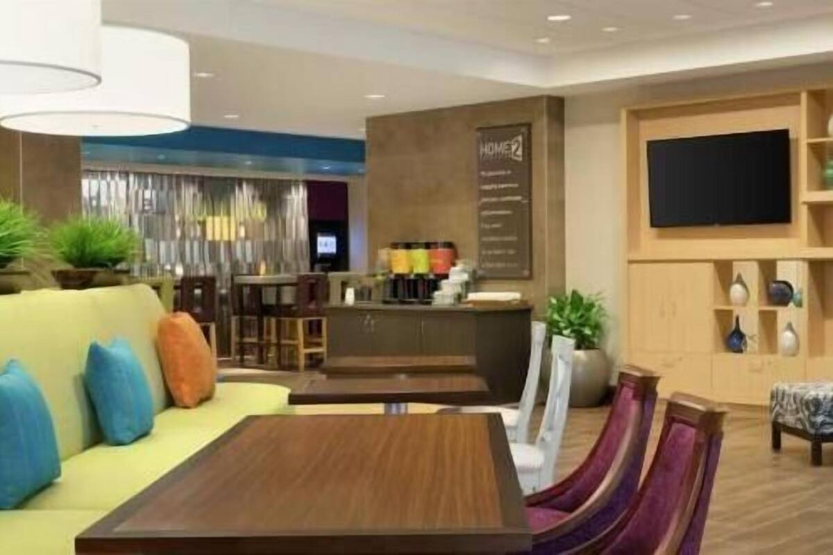 A modern hotel lobby with a sofa, a wooden table, a bar area, and a TV screen.