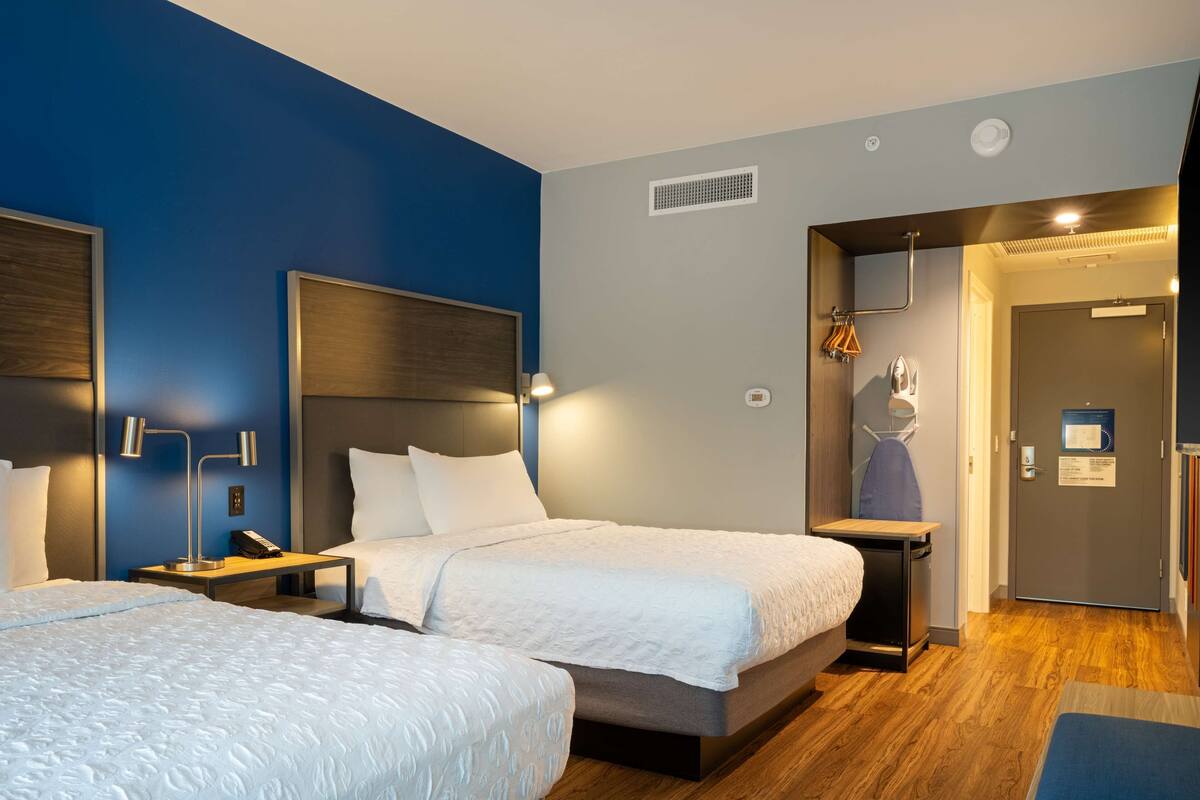 Ein Hotelzimmer mit zwei Betten, Holzboden, blauer Akzentwand und einer Tür zum Badezimmer.