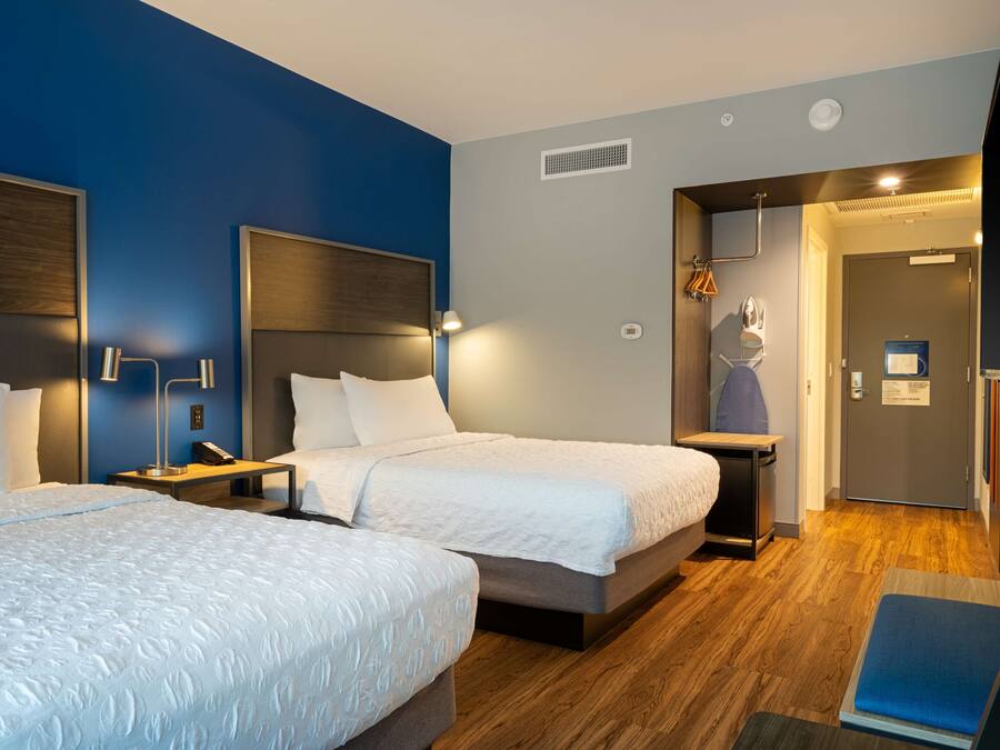 A hotel room with two beds, a wooden floor, a blue accent wall, and a door leading to a bathroom.